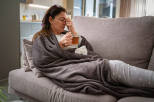 mulher, deitada no sofá, com uma coberta cinza a envolvendo, enquanto segura uma xícara de chá em uma mão e aperta seu nariz com a outra mão, com os olhos fechados e em uma expressão de dor