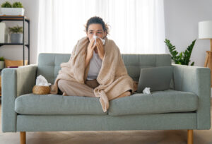 mulher sentada em seu sofá cinza, enrolada em uma coberta, enquaanto assoaa seu nariz com um papel para aliviar a coriza da gripe