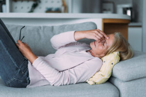 mulher branca deitada em seu sofá com uma mão apertando o meio dos olhos para indicar cansaço